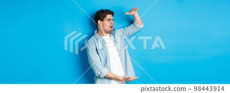 Excited handsome man showing big size object and looking amazed, standing over blue background Excited handsome man showing big size object and looking amazed, standing over blue background 98443914