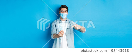 Concept of coronavirus, quarantine and social distancing. Young man pointing at medical masks, preventing measures from covid-19, standing over blue background 98443918