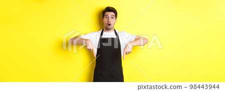 Surprised seller in black apron pointing fingers down, looking amazed at camera, standing over yellow background Surprised seller in black apron pointing fingers down, looking amazed at camera, standing over yellow background 98443944