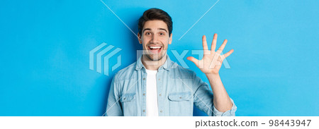 Close-up of handsome man smiling, showing fingers number five, standing over blue background Close-up of handsome man smiling, showing fingers number five, standing over blue background 98443947
