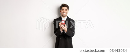 Image of handsome boyfriend in black suit making a proposal, asking to marry him and showing wedding ring, standing over white background Image of handsome boyfriend in black suit making a proposal, asking to marry him and showing wedding ring, standing over white background 98443984