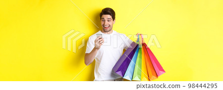 Excited man holding shopping bags and looking happy at mobile phone screen, standing over yellow background 98444295