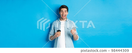 Happy young man drinking coffee and using mobile phone, looking excited, standing against blue background 98444309