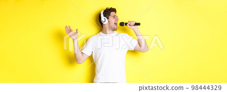 Passionate guy in headphones holding microphone, singing karaoke song, standing over yellow background in white clothes Passionate guy in headphones holding microphone, singing karaoke song, standing over yellow background in white clothes 98444329