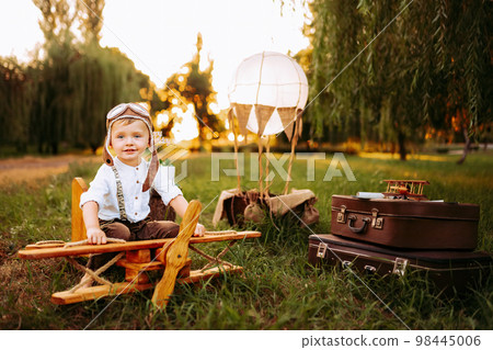 Cute little pilot boy in vintage aviator hat looking at camera while sitting on big toy wooden plane Cute little pilot boy in vintage aviator hat looking at camera while sitting on big toy wooden plane 98445006
