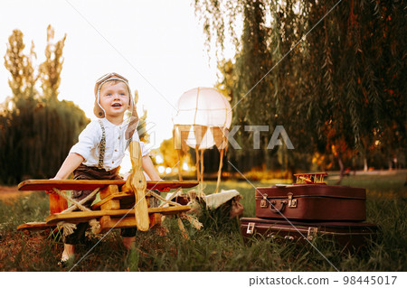 Dreaming little pilot boy in vintage aviator hat looking forward while sitting on big toy wood plane Dreaming little pilot boy in vintage aviator hat looking forward while sitting on big toy wood plane 98445017