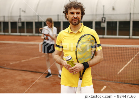 Portrait of male tennis player and female behind. Man standing on a court and looking at camera. Man wearing yellow t-shirt and white shorts. 98447166