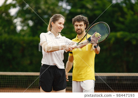Man and woman tennis players standing on open summer court with rackets in hands. Man teaches woman how to hold a racket. Man wearing yellow t-shirt and woman white one. Man and woman tennis players standing on open summer court with rackets in hands. Man teaches woman how to hold a racket. Man wearing yellow t-shirt and woman white one. 98447193