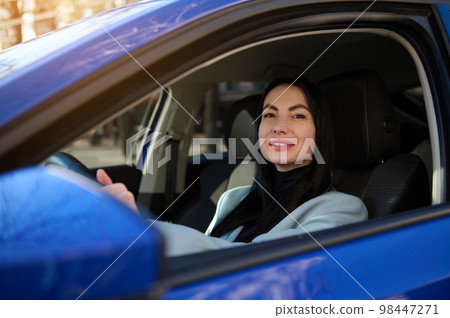 Close up portrait of Caucasian confident 40-44 year old beautiful business woman, driver putting hands on the steering wheel, smiling looking at camera while driving car. People and Urban Lifestyles Close up portrait of Caucasian confident 40-44 year old beautiful business woman, driver putting hands on the steering wheel, smiling looking at camera while driving car. People and Urban Lifestyles 98447271
