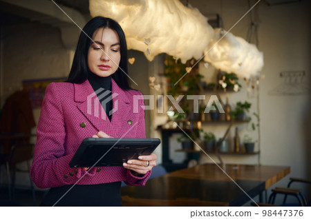 Charming middle-aged dark-haired Caucasian woman in elegant stylish casual wear, purposeful restaurateur, CEO, successful manager, business lady working on tablet in the modern restaurant interior 98447376