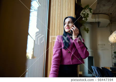 Confident middle-aged Caucasian woman in stylish formal wear, sitting on the window sill, talking on mobile phone, looking through the window in conference room. People. Communication. Business Confident middle-aged Caucasian woman in stylish formal wear, sitting on the window sill, talking on mobile phone, looking through the window in conference room. People. Communication. Business 98447394