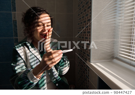 Middle aged multiracial happy woman feeling happiness while holding a positive inkjet pregnancy test, standing by the window while shadows from window blinds reflect on her face in the home bathroom Middle aged multiracial happy woman feeling happiness while holding a positive inkjet pregnancy test, standing by the window while shadows from window blinds reflect on her face in the home bathroom 98447427
