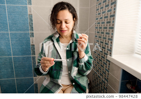 Charming multi-ethnic woman with nervous feelings, sitting on the toilet bowl, taking pregnancy test and nervously awaiting the result, smiling from happiness while rejoicing at the positive result Charming multi-ethnic woman with nervous feelings, sitting on the toilet bowl, taking pregnancy test and nervously awaiting the result, smiling from happiness while rejoicing at the positive result 98447428