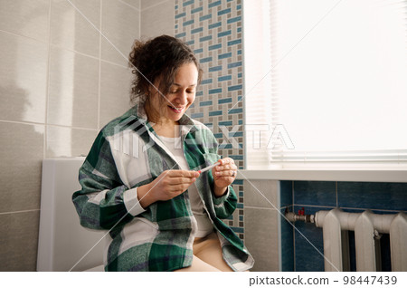 African American middle-aged positive woman, sitting on toilet bowl, taking pregnancy test in the home bathroom 98447439