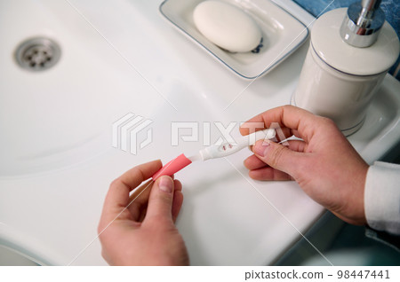 Top view woman hands holding an inkjet pregnancy test with positive result in action over white wash basin in the home bathroom. Finally pregnant 98447441