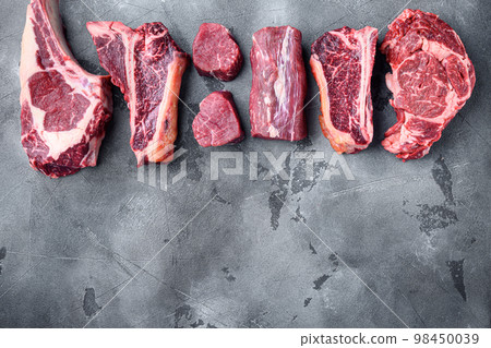 Variety of raw beef meat steaks for grilling with seasoning and utensils, tomahawk, t bone, club steak, rib eye and tenderloin cuts, on gray stone background, top view flat lay, with copy space for 98450039
