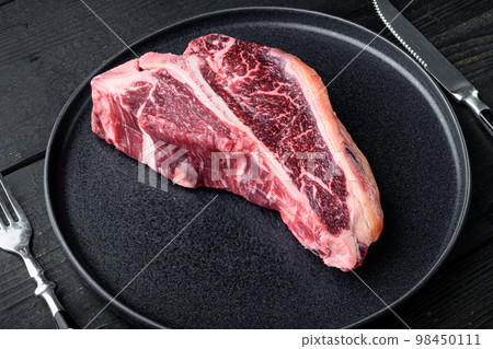 Raw fresh beef t-bone steak drya aged cut, on plate, on black wooden table background, with copy space for text 98450111