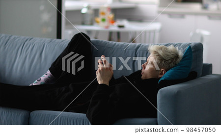 Blond young man is relaxing on grey sofa, using his gadget, smartphone, texting, editing, browsing, looking at screen, smiling, spending weekend at home. 98457050