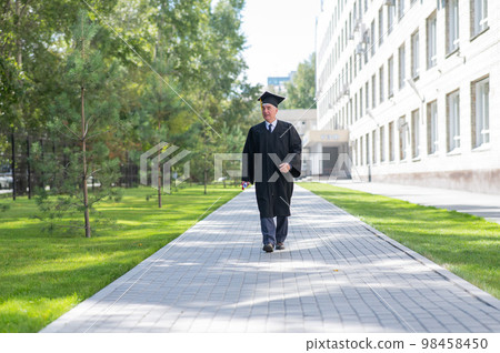 An old man in a graduation gown walks outdoors and holds a diploma. An old man in a graduation gown walks outdoors and holds a diploma. 98458450