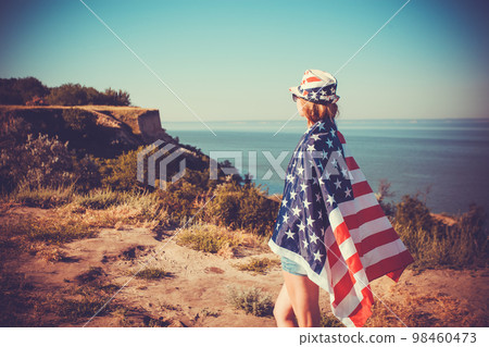 Beautiful young girl holding an American flag on the wind in a meadow. Summer landscape against the blue sky. Beautiful young girl holding an American flag on the wind in a meadow. Summer landscape against the blue sky. 98460473