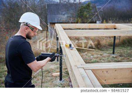 Man building frame house on pile foundation. Male worker drilling hole by electric drill in wooden frame of future house. Builder wearing white helmet and protective goggles. Carpentry concept. 98461527