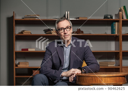 Happy grey haired man in blue suit and glasses is sitting in livingroom at home 98462625