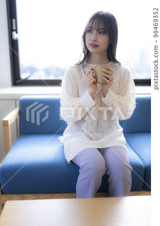 Woman relaxing on the sofa in the office with a cup of coffee. An expression looking somewhere. Cityscape through the window. Woman relaxing on the sofa in the office with a cup of coffee. An expression looking somewhere. Cityscape through the window. 98469352