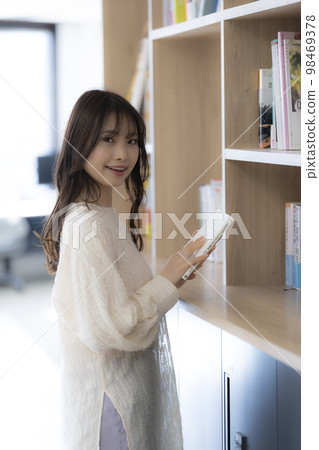 年輕女子在辦公室書架上找書。有一條視線。 98469378