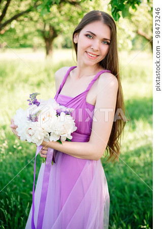 Portrait of a young woman 20s 30s in a light purple dress with a bouquet of peonies in her hands. Graduation, bridesmaid posing in the garden. Portrait of a young woman 20s 30s in a light purple dress with a bouquet of peonies in her hands. Graduation, bridesmaid posing in the garden. 98473246