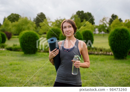 A young athletic girl in a gray tracksuit for fitness is going to do yoga in a green park, in her hands a bottle of water and a yoga mat. A young athletic girl in a gray tracksuit for fitness is going to do yoga in a green park, in her hands a bottle of water and a yoga mat. 98473260
