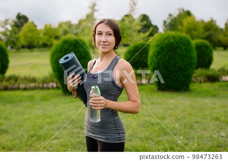 A young athletic girl in a gray tracksuit for fitness is going to do yoga in a green park, in her hands a bottle of water and a yoga mat.  98473263