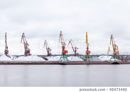 River port in winter. Port cranes on the bank of the river. Embankment of the river port, a barge at the quay wall. Large port cranes on the river. Extraction and transportation of sand other cargo 98473490