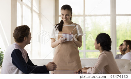 Pretty young waitress wearing uniform apron, serving diverse couple Pretty young waitress wearing uniform apron, serving diverse couple 98474180