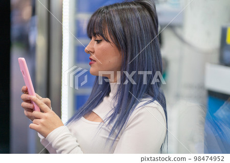 Concentrated stylish young female with blue hair in trendy outfit standing near glass window on street and browsing internet via smartphone 98474952
