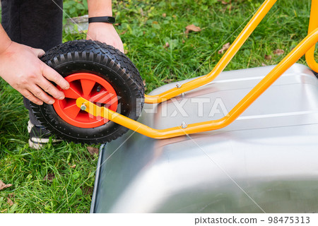 There is a metal wheelbarrow on the street, a man spins a new wheelbarrow for work, tightens bolts and nuts, hangs a wheel. There is a metal wheelbarrow on the street, a man spins a new wheelbarrow for work, tightens bolts and nuts, hangs a wheel. 98475313