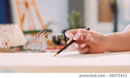 Drawing class. Art hobby. Creative occupation. Closeup of female artist hand sketching artwork with pencil on white canvas at workplace with free space. Drawing class. Art hobby. Creative occupation. Closeup of female artist hand sketching artwork with pencil on white canvas at workplace with free space. 98475413