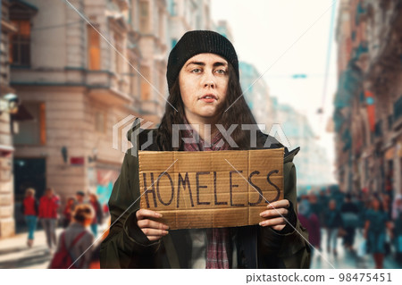 A beggar senior caucasian woman holds a cardboard sign with the inscription homeless. Unfocused background with street and people. The concept of helping vagrants 98475451