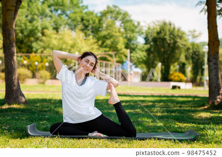 Yoga. A young pretty woman in sportswear performs an exercise sitting on a Mat in a Park on the grass. The concept of a healthy lifestyle 98475452