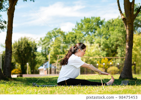Yoga. A young woman in sports clothes, performs a tilt, sitting on a mat, in a Park on the grass. Side view. The concept of sport for a healthy lifestyle 98475453