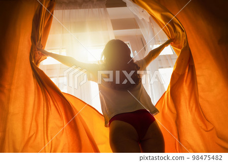New day and sunlight morning. Silhouette of woman opening orange curtains at window. Bottom view. Concept of freedom, hope and happiness 98475482