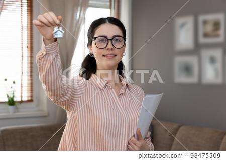 Portrait of woman realtor wearing glasses showing keys. Cozy interior in background. Concept of purchase real estate 98475509