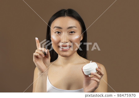 Happy young japanese lady holding jar of cream and smiling at camera, posing isolated on brown background, studio shot Happy young japanese lady holding jar of cream and smiling at camera, posing isolated on brown background, studio shot 98475970