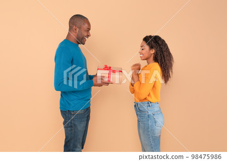 Loving black man giving gift to her young wife, standing over peach studio background, free space, side view Loving black man giving gift to her young wife, standing over peach studio background, free space, side view 98475986