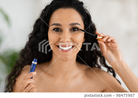 Portrait of smiling muslim millennial curly woman applying mascara on eyelashes looking in camera Portrait of smiling muslim millennial curly woman applying mascara on eyelashes looking in camera 98475987