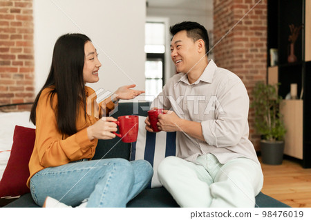 Happy asian spouses sitting on sofa with cups of hot drink and talking, resting in living room interior 98476019