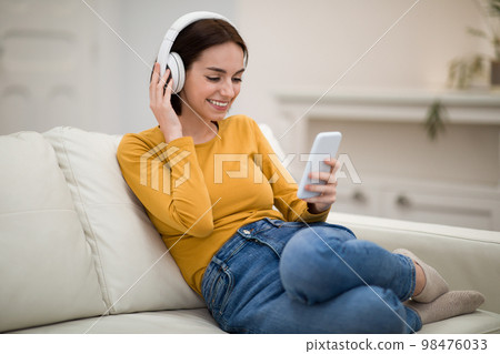 Positive young lady using headphones and cell phone at home Positive young lady using headphones and cell phone at home 98476033