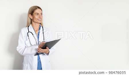 Medical Offer. Smiling female doctor in uniform holding clipboard and looking aside Medical Offer. Smiling female doctor in uniform holding clipboard and looking aside 98476035