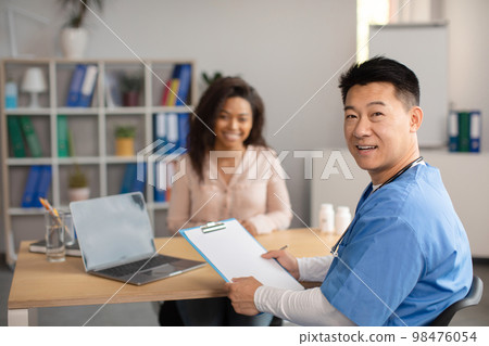 Smiling middle aged korean doctor in uniform consults young african american lady in clinic office interior Smiling middle aged korean doctor in uniform consults young african american lady in clinic office interior 98476054