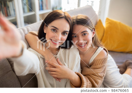 Happy european young women take selfie together, sit on sofa, have fun in free time in living room interior Happy european young women take selfie together, sit on sofa, have fun in free time in living room interior 98476065