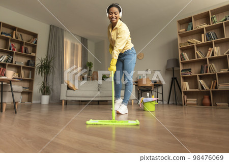 Young black woman washing floor in living room using mop and listening music in headphones, enjoying cleaning routine Young black woman washing floor in living room using mop and listening music in headphones, enjoying cleaning routine 98476069